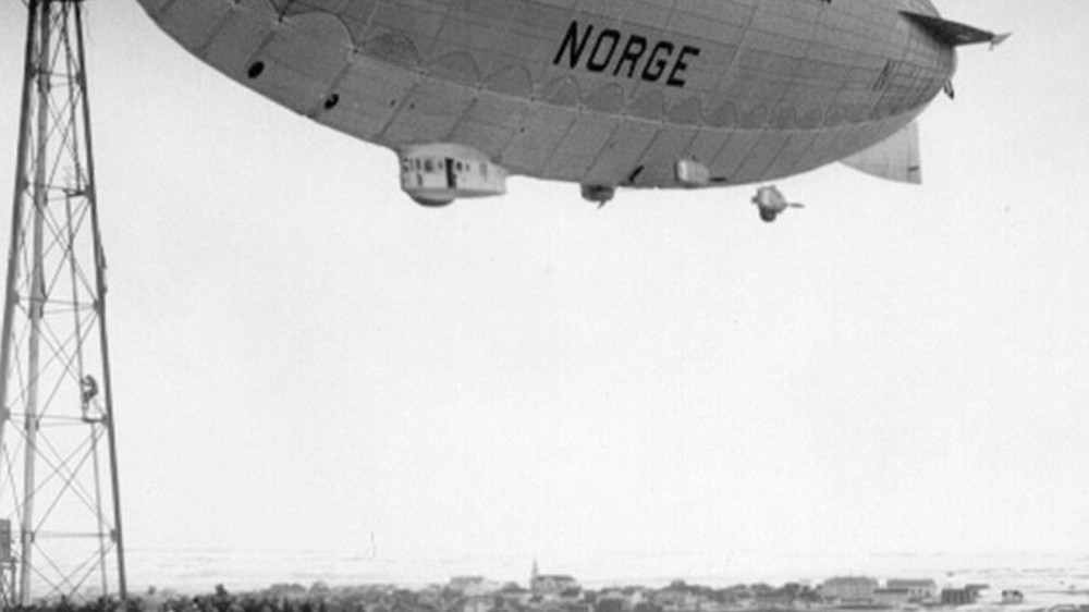 Luftskipet "Norge" svever over bakken ved siden av en høy mast, med mennesker som ser på fra bakken. Foto.