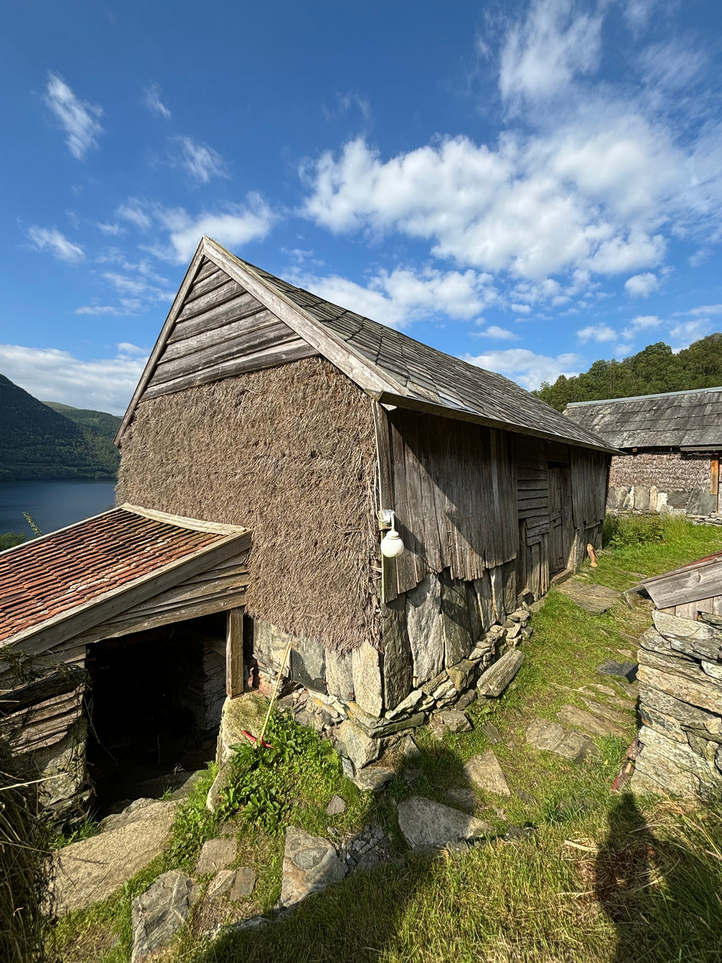 Løe fra 1878 med skifertak og kledning av tre og halm. Beliggende på gårdstun med fjordlandskap og himmel i bakgrunnen.