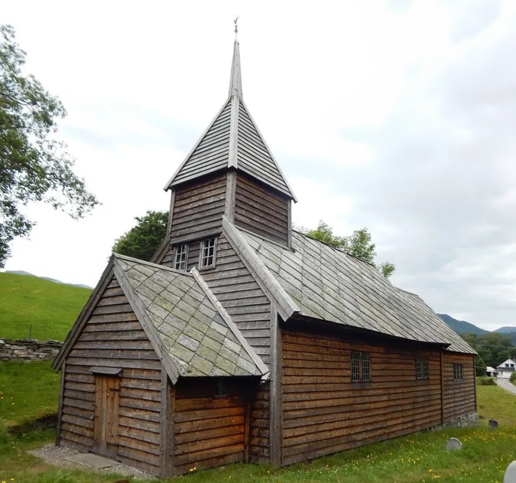 En gammel trekirke med tårn og våpenhus, omgitt av grønt gress og trær. Foto.