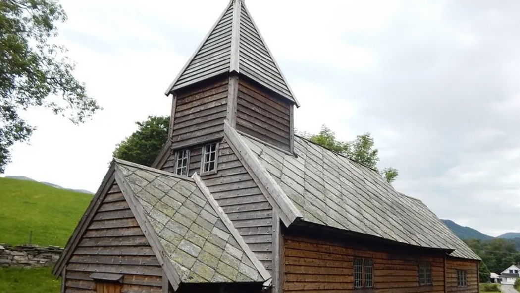 En gammel trekirke med tårn og våpenhus, omgitt av grønt gress og trær. Foto.