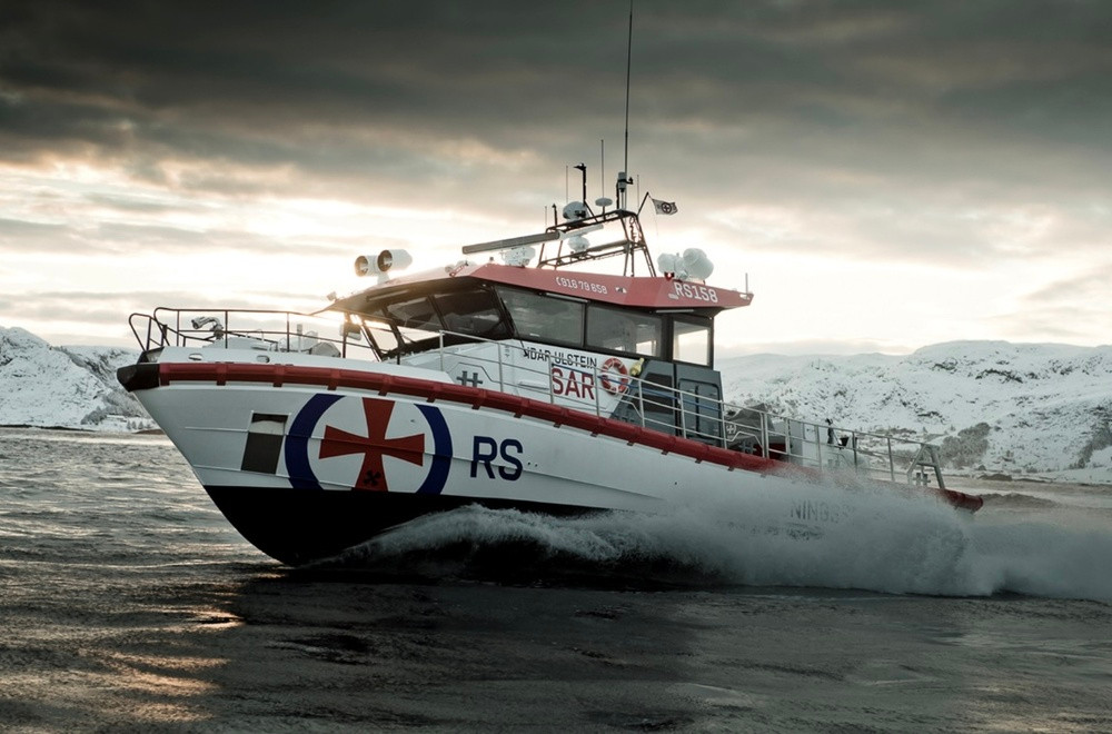 En hvit og rød redningsskøyte i fart på havet med fjell i bakgrunnen under en dramatisk himmel. Foto.