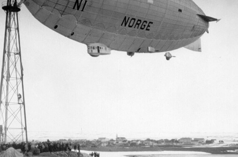 Luftskipet "Norge" svever over bakken ved siden av en høy mast, med mennesker som ser på fra bakken. Foto.